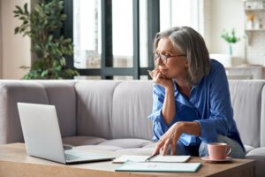 An older woman uses research on her laptop while holding a notepad.